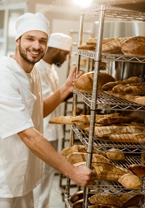 Preview: baker near shelves with fresh bread loaves at baking manufacture