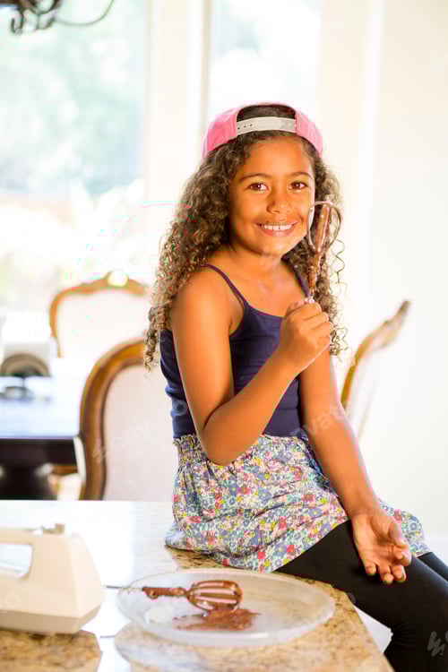 Visualização: Garota sentada no balcão da cozinha segurando um batedor coberto com mistura de bolo olhando para a câmera sorrindo