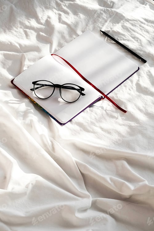 Preview: Blank Notebook, Pencil, Eyeglasses Rest on White Bedding in Sunlight with Shadows.