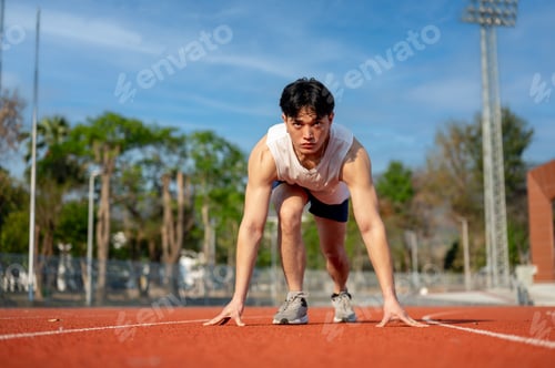 Preview: Asian man in white tank top crouching in a set position ready to run on racetrack. Outdoor exercise.