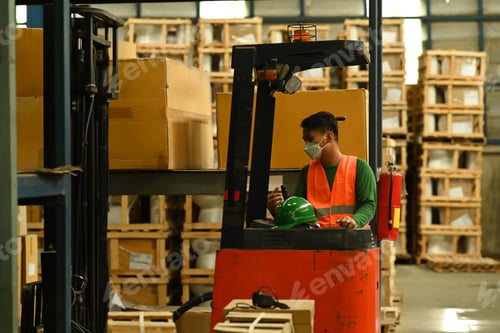Preview: Male warehouse worker moving cardboard boxes of goods with forklift between rows of tall shelves.