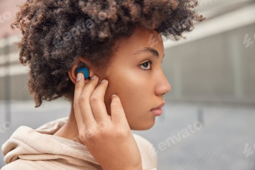 Preview: Sideways shot of serious curly woman puts on wireless earbuds listens music favorite playlist during