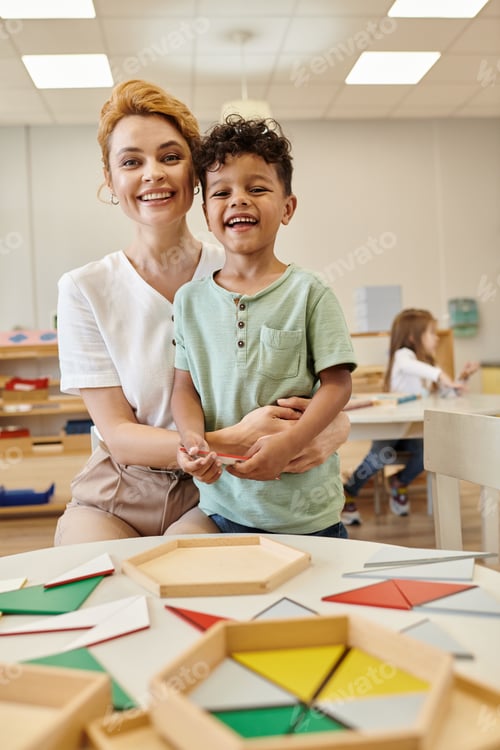 Preview: smiling teacher hugging african american kid near didactic materials in montessori school