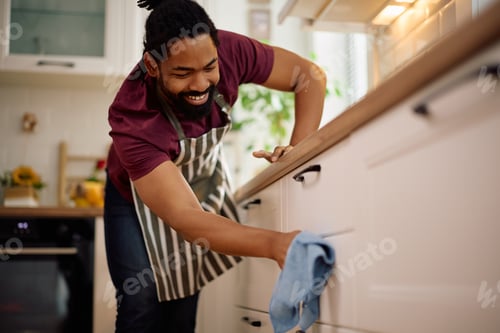 Preview: Happy black man cleaning kitchen counters at home.