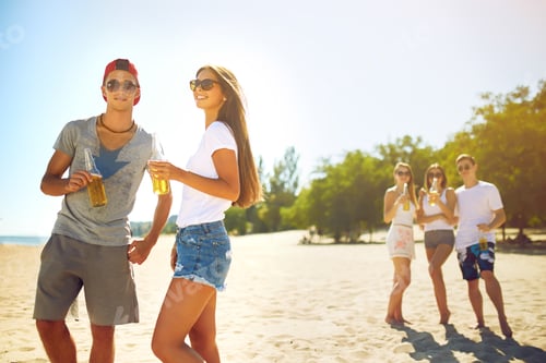 Preview: Smiling young couple clinking glasses with beer while people barbecuing in the background.
