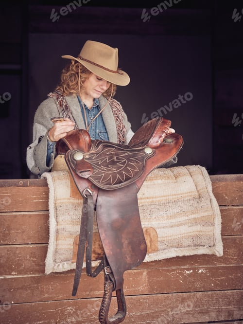 Preview: Lady in cowgirl hat placing saddle on carpet in stall