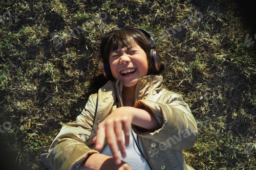 Preview: Funny little asian girl wearing headphones lying on grass laughing having fun listening to music in