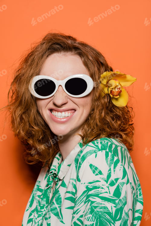 Preview: Positive woman with orchid flower in red hair wearing sunglasses and blouse with floral pattern