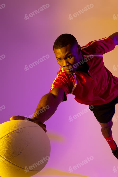 Preview: African american male rugby player jumping with rugby ball over pink lighting