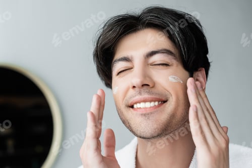 Preview: happy man with closed eyes applying face cream in bathroom