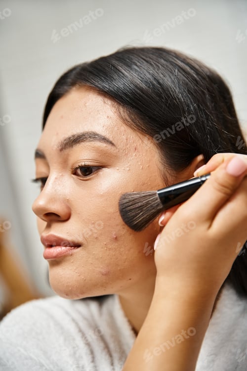 Preview: brunette and young asian woman applying face powder over acne-prone skin with cosmetic brush