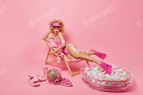 Preview: Surprised curly haired woman applies suncream on skin to protect from sunburn wears swimsuit sunglas