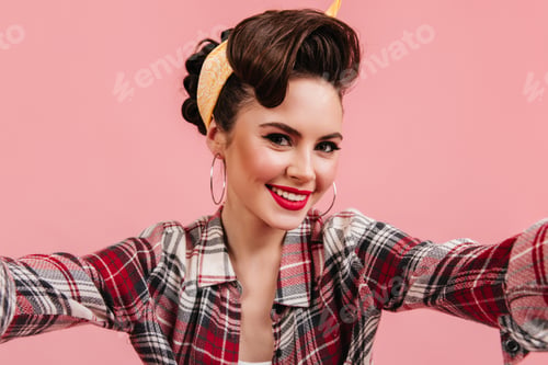 Preview: Lovely young woman in checkered shirt smiling on pink background. Emotional pinup girl taking selfi