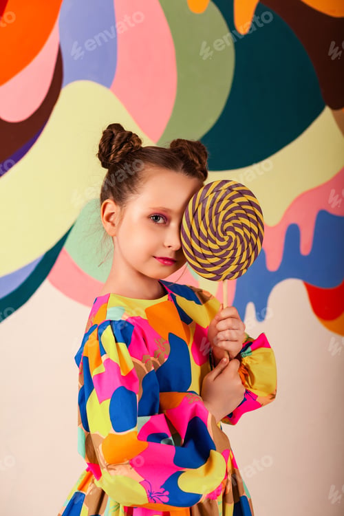 Preview: Adorable little girl covering eye round lollipop at multi colored wall background