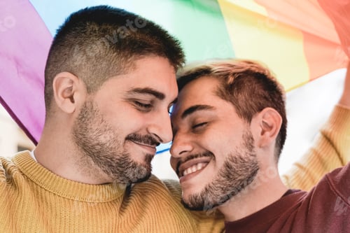Preview: Young gay men couple having tender moment under rainbow LGBT flag outdoor - LGBTQ+ love concept