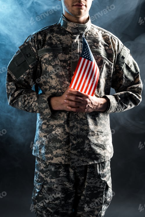 Preview: cropped view of man in camouflage uniform holding american flag on black with smoke