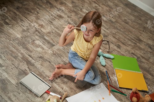 Preview: Cute little caucasian girl holding lollipop and smiling while sitting on the wooden floor in