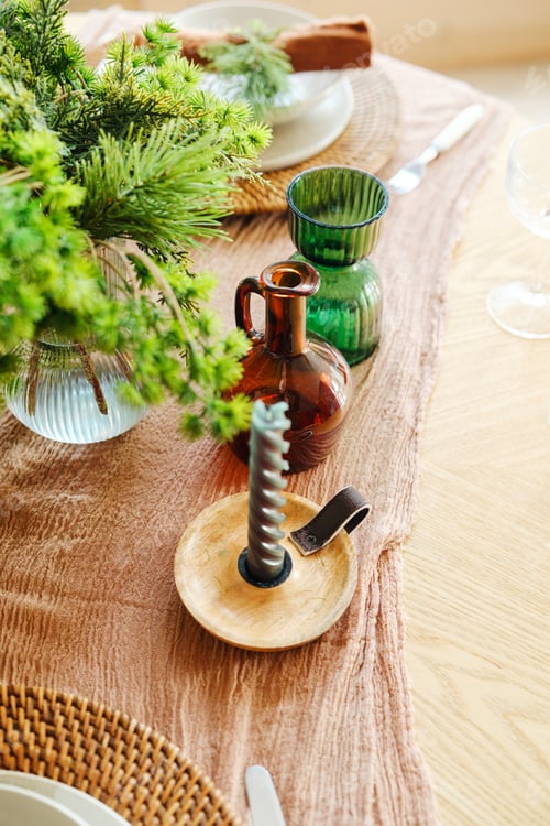 Preview: Table setting with evergreen branches, amber bottle, green vase, and candlestick on wood