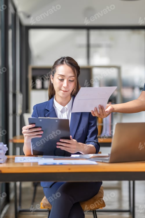 Preview: Brown haired wearing light blue jacket smiling Asian woman work with document laptop in office,