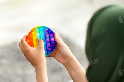 Preview: Cropped view of Muslim teenage girl in hijab playing with antistress POP IT toy at home, closeup of