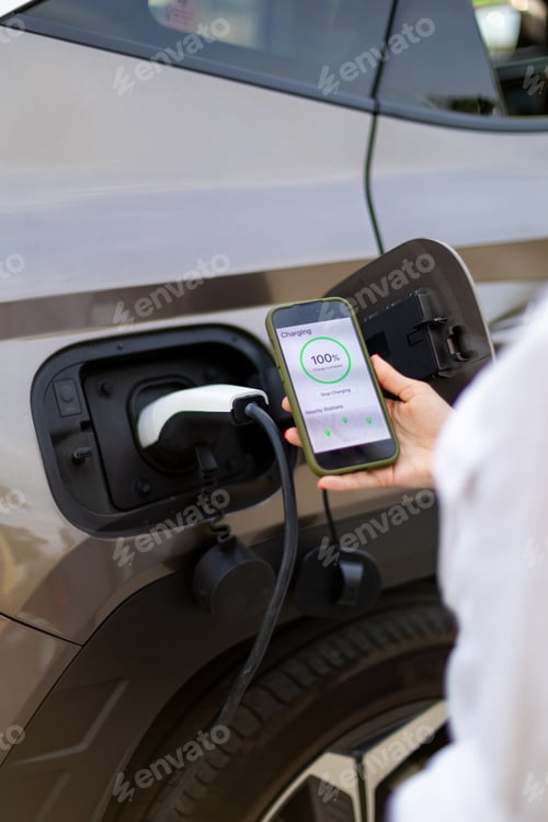 Preview: Businesswoman using smartphone app while charging electric car at public station
