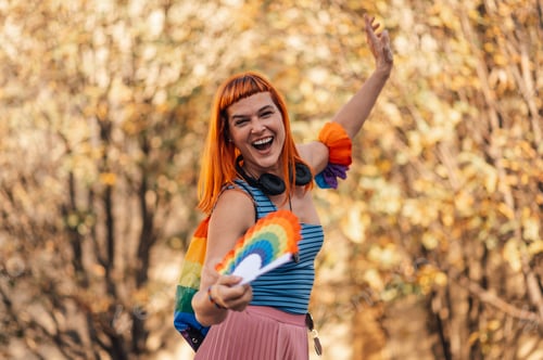Preview: Young redhead woman celebrating gay pride holding a rainbow fan