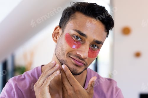 Preview: Smiling biracial man with eye masks inspecting his face in bathroom mirror