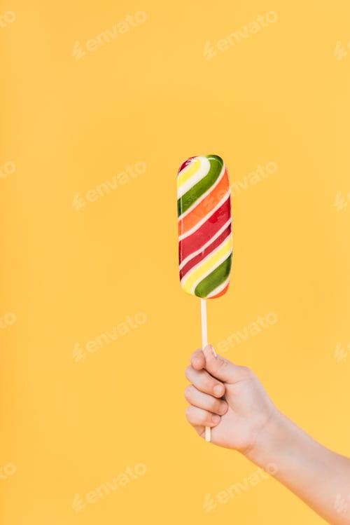 Preview: partial view of kid holding lollipop in hand isolated on yellow