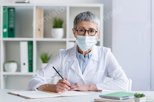 Preview: Mature doctor in medical mask holding pen near notebooks on blurred foreground