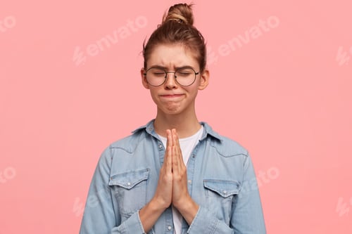 Preview: Sorrowful Caucasian female keeps palms in praying gesture, lips pressed and eyes shut, wears denim s