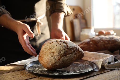 Preview: Man holding loaf of fresh bread at wooden table indoors, closeup