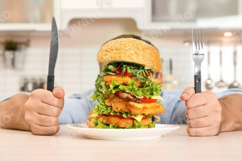 Preview: Young hungry man with cutlery eating huge burger at table