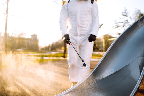Preview: Man wearing protective suit disinfecting the playground in the sun with spray. Covid -19. Cleaning.