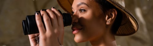 Preview: young archaeologist in safari hat looking through binoculars in desert, banner