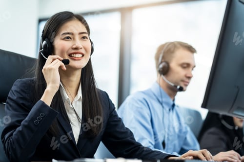 Preview: Asian female holding headset microphone smiling when talking to customers while colleagues working.
