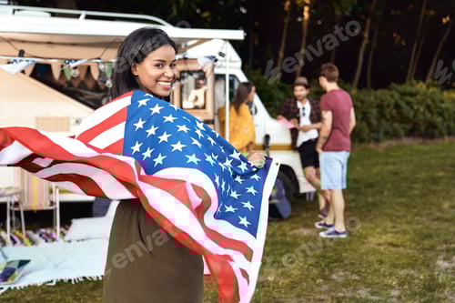 Preview: Young mixed race woman covered with USA flag and her friends celebrating 4th of July on the camping