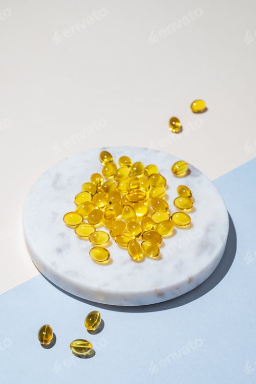 Preview: Yellow softgel vitamins capsules on a white marble plate on a blue background with shadow.