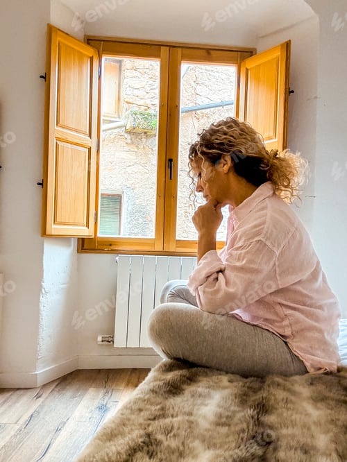 Preview: Woman sitting by a sunny window in thoughtful pose at home reflecting and relaxing in a cozy room