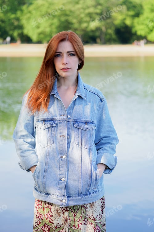 Preview: young woman wearing denim jacket over summer dress standing by lake - authentic real people concept