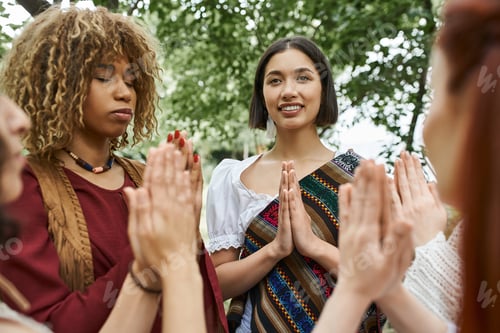 Vista previa: mujer joven positiva vestida de boho meditando cerca de amigos multiétnicos al aire libre en un centro de retiros