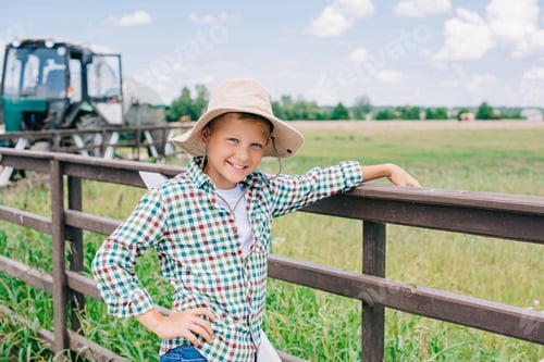 Preview: cheerful child in panama hat leaning at fence and smiling at camera on farm