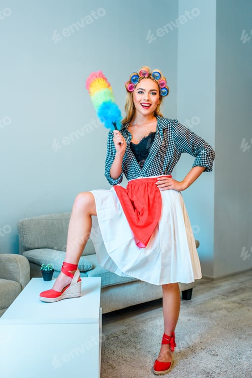 Preview: beautiful blonde smiling housewife in curlers cleaning house with duster