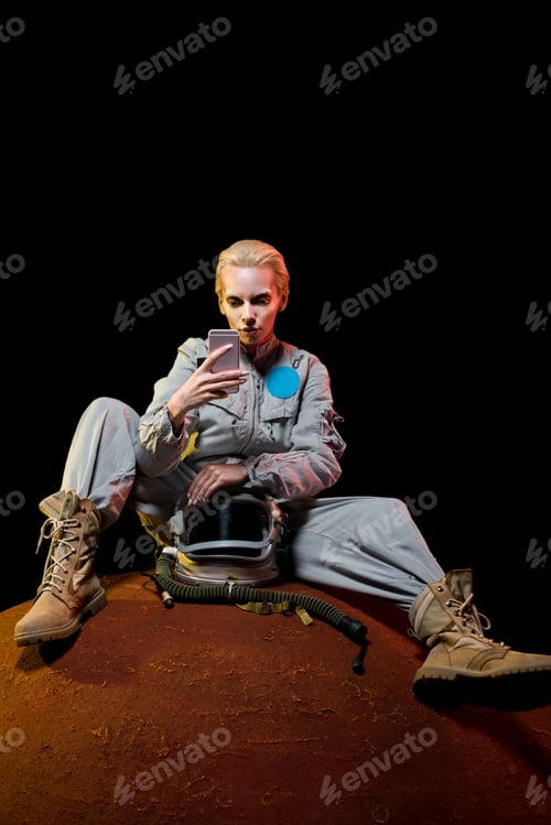 Preview: astronaut in spacesuit with helmet sitting on planet and using smartphone