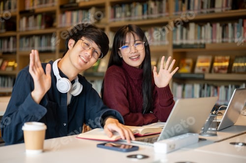 Preview: Smiling glasses asian man student and a woman friend waving a hand while sitting at library's table.