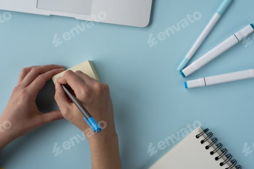 Preview: High angle shot of a person writing on sticky notes and a journal, pens, and laptop on the table