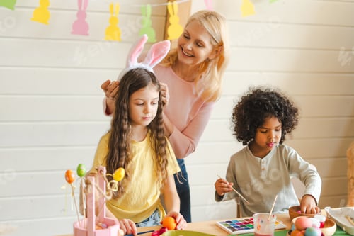 Preview: Grandmother putting bunny ears on girl, multi ethnic children decorating Easter eggs