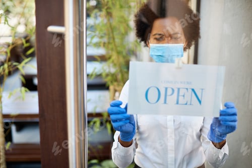 Preview: Black waitress with protective face mask and gloves hanging open sign at entrance door.