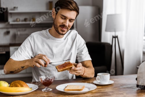 Preview: happy young man applying jam onto toast at home