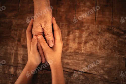 Preview: Two hands, one aged and one youthful, gently clasping each other on a wooden surface