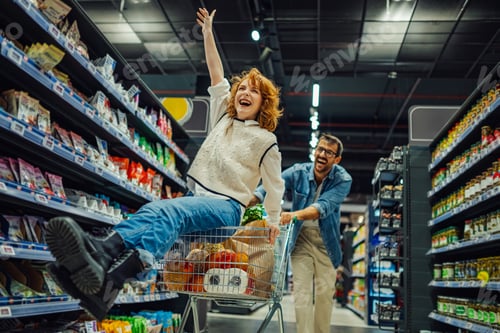 Preview: Happy couple having fun while grocery shopping in supermarket, woman sitting in shopping cart